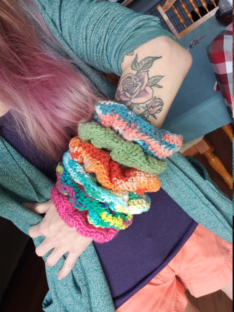 May include: A stack of colorful crocheted bracelets in various shades of pink, green, orange, blue, and white. The bracelets are made with a textured stitch and are stacked on top of each other.