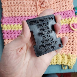 Op de afbeelding: Een zwart garen gewicht meetinstrument met tekst die garen gewichten aangeeft van Super Bulky tot Lace. De achtergrond toont een kleurrijk haakproject in tinten perzik, roze en geel, met een bol garen.