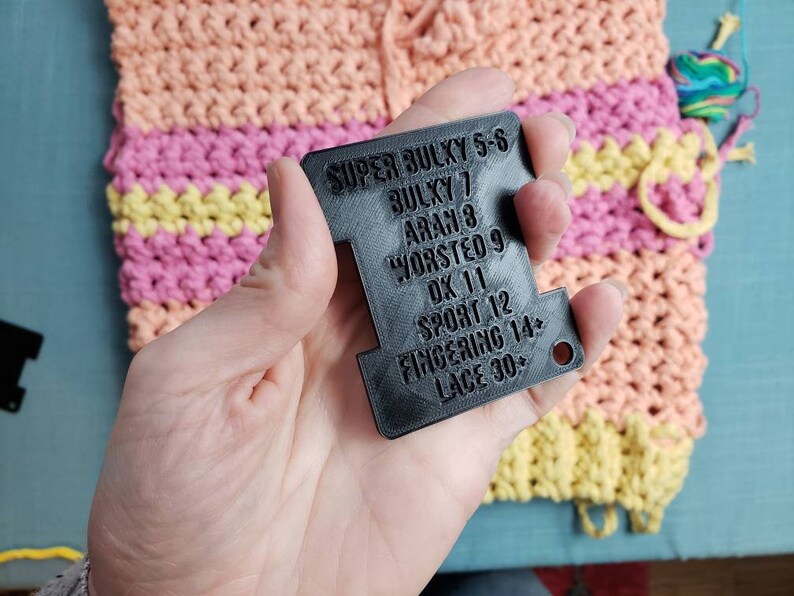 Op de afbeelding: Een zwart garen gewicht meetinstrument met tekst die garen gewichten aangeeft van Super Bulky tot Lace. De achtergrond toont een kleurrijk haakproject in tinten perzik, roze en geel, met een bol garen.