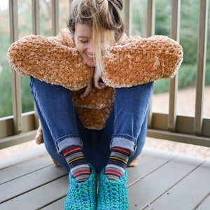 May include: A person wearing a brown fuzzy jacket and blue jeans sits on a wooden deck. They are wearing colorful striped socks and blue crocheted slippers.