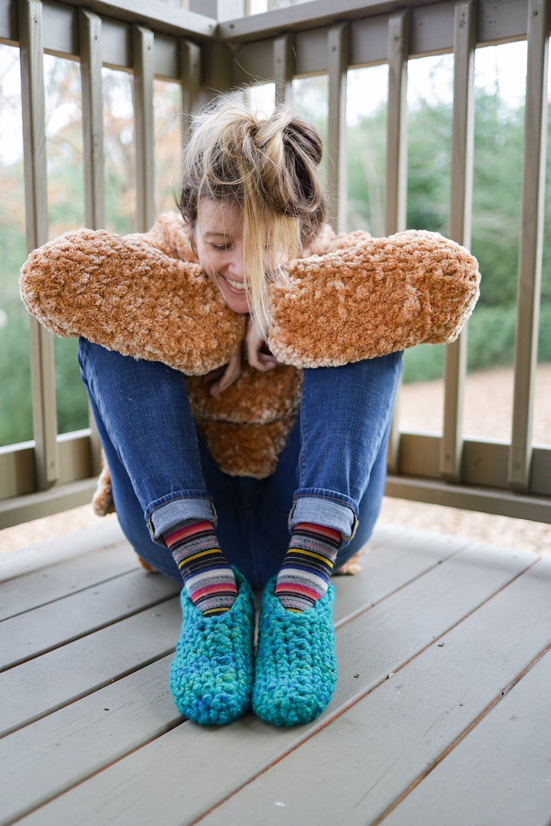 May include: A person wearing a brown fuzzy jacket and blue jeans sits on a wooden deck. They are wearing colorful striped socks and blue crocheted slippers.