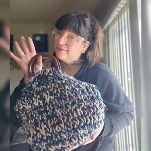 May include: A chunky knit handbag with a dark brown wooden handle. The bag features a mix of navy blue, cream, and purple yarn. The bag is held up by a person.