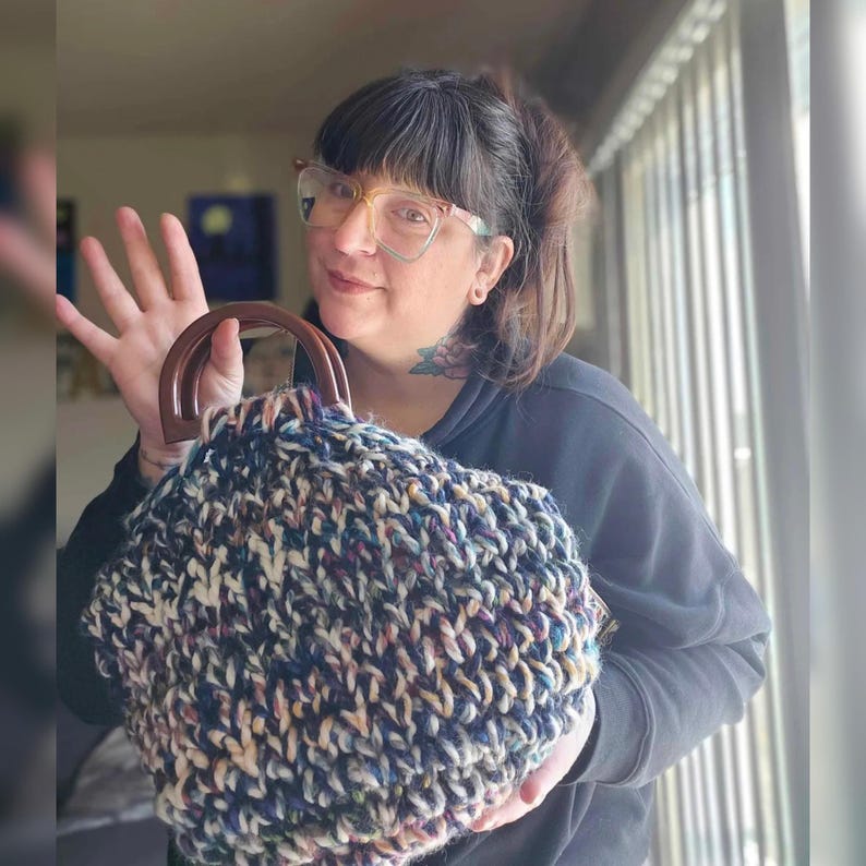 May include: A chunky knit handbag with a dark brown wooden handle. The bag features a mix of navy blue, cream, and purple yarn. The bag is held up by a person.