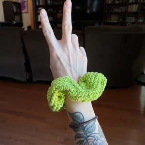 May include: A bright green crocheted hair scrunchie. The scrunchie is on a person's wrist, and the person has a tattoo of a rose on their arm.