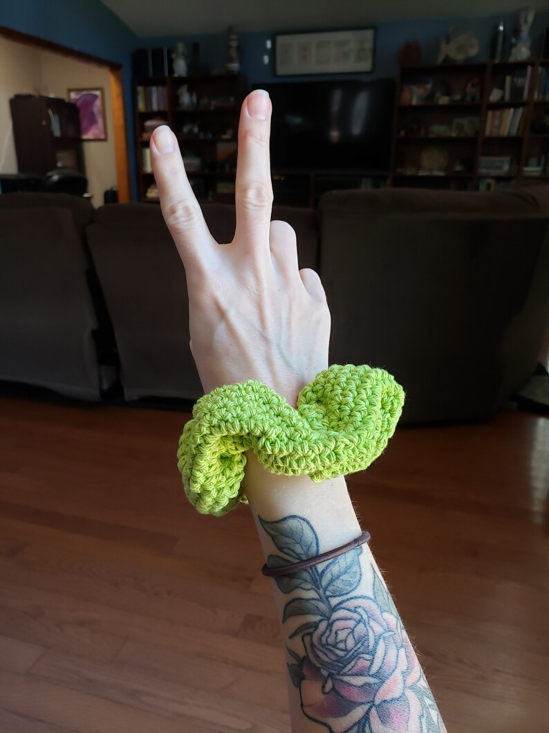 May include: A bright green crocheted hair scrunchie. The scrunchie is on a person's wrist, and the person has a tattoo of a rose on their arm.