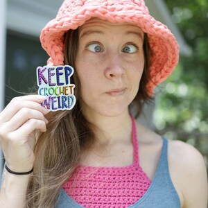 May include: A woman wearing a pink crocheted sun hat and a blue tank top with a white crocheted halter top. She is holding a sticker that says "Keep Crochet Weird" in rainbow letters.