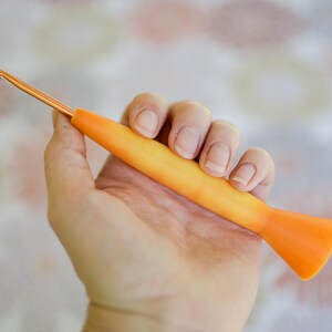 May include: An orange and yellow crochet hook with a metallic gold hook end. The hook is held in a hand against a soft, floral patterned background. The hook is a crafting tool for crochet projects.