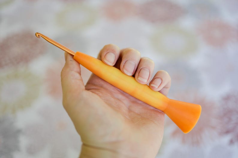 May include: An orange and yellow crochet hook with a metallic gold hook end. The hook is held in a hand against a soft, floral patterned background. The hook is a crafting tool for crochet projects.