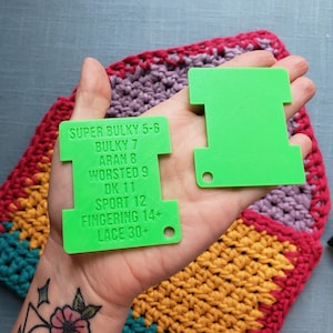 Op de afbeelding: Twee felgroene garenmeters worden in een hand gehouden, met een kleurrijk gehaakt item op de achtergrond. E&eacute;n tool heeft tekst met garendiktes: Super Bulky, Bulky, Aran, Worsted, DK, Sport, Fingering en Lace.