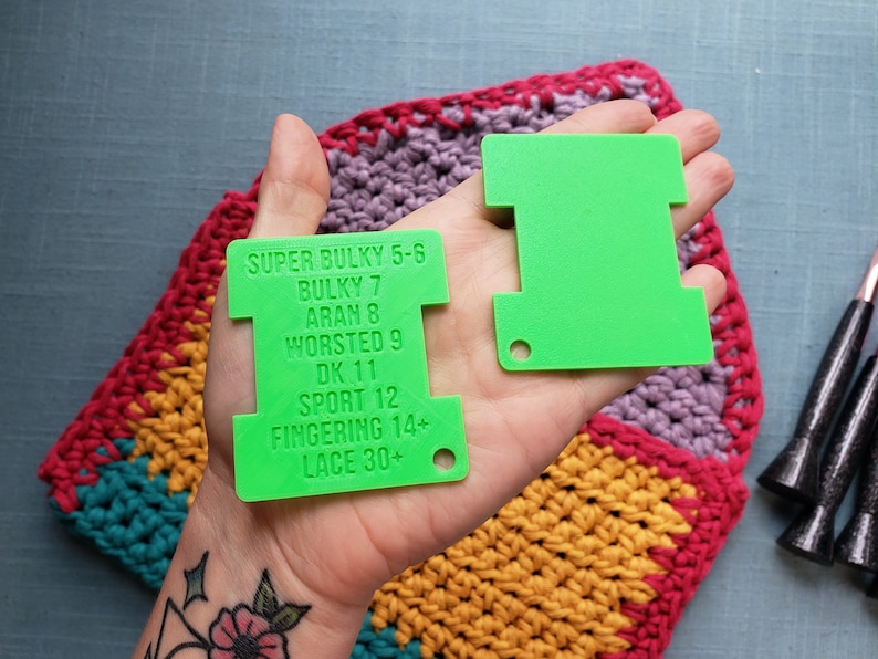 Op de afbeelding: Twee felgroene garenmeters worden in een hand gehouden, met een kleurrijk gehaakt item op de achtergrond. E&eacute;n tool heeft tekst met garendiktes: Super Bulky, Bulky, Aran, Worsted, DK, Sport, Fingering en Lace.