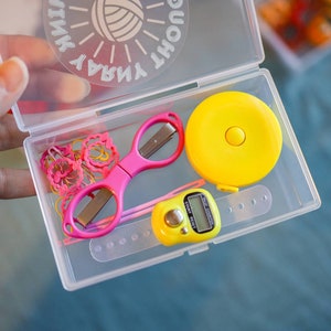 Pode incluir: Uma caixa de armazenamento de pl&aacute;stico transparente com uma fita m&eacute;trica amarela, tesoura rosa, um contador de pontos amarelo e el&aacute;sticos de cabelo rosa. A tampa da caixa tem o texto "Yarn Thought Sink" impresso.