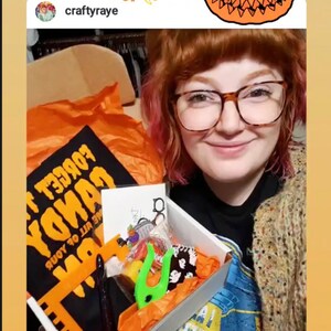 Puede incluir: Una mujer con gafas y un su&eacute;ter sostiene una caja llena de art&iacute;culos con tem&aacute;tica de Halloween. La caja contiene una camiseta negra con el texto "Forget Your Candy" en letras naranjas, una mano de pl&aacute;stico verde y otras golosinas de Halloween. El texto "craftyraye I received my Halloween mystery box from @pinksheepdesign &... more" es visible en la parte inferior de la imagen.
