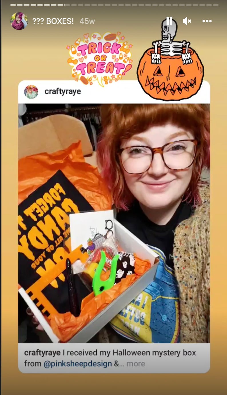 Puede incluir: Una mujer con gafas y un su&eacute;ter sostiene una caja llena de art&iacute;culos con tem&aacute;tica de Halloween. La caja contiene una camiseta negra con el texto "Forget Your Candy" en letras naranjas, una mano de pl&aacute;stico verde y otras golosinas de Halloween. El texto "craftyraye I received my Halloween mystery box from @pinksheepdesign &... more" es visible en la parte inferior de la imagen.