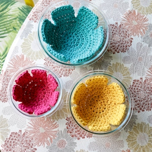 May include: Three crocheted bowl liners in a floral pattern tablecloth. The liners are teal, pink, and yellow.