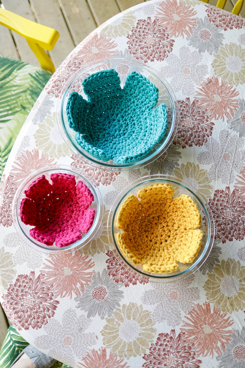 May include: Three crocheted bowl liners in a floral pattern tablecloth. The liners are teal, pink, and yellow.