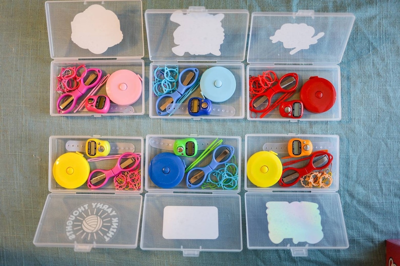 Pode incluir: Seis recipientes de pl&aacute;stico transparente com tampas, cada um contendo um conjunto de ferramentas de costura e tric&ocirc; coloridas. Os recipientes s&atilde;o etiquetados com as palavras "Think Yarn" e "Yarn Thought".