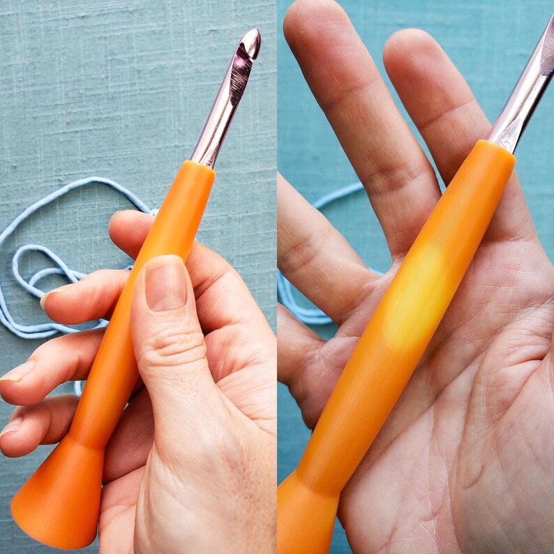 May include: An orange crochet hook with a silver metal hook end. The hook has a smooth, ergonomic handle. The image shows the hook being held in a hand, with blue yarn in the background. The hook is designed for yarn crafts.
