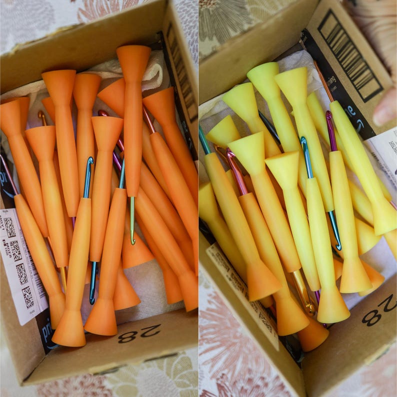 May include: A cardboard box containing crochet hooks in orange and yellow. The hooks have metal tips in various colours, such as blue, pink, and green. The handles are a solid colour, and the hooks are neatly arranged.