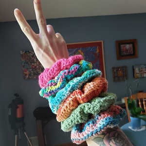 May include: A stack of six colorful crocheted hair scrunchies in pink, green, blue, orange, and teal.