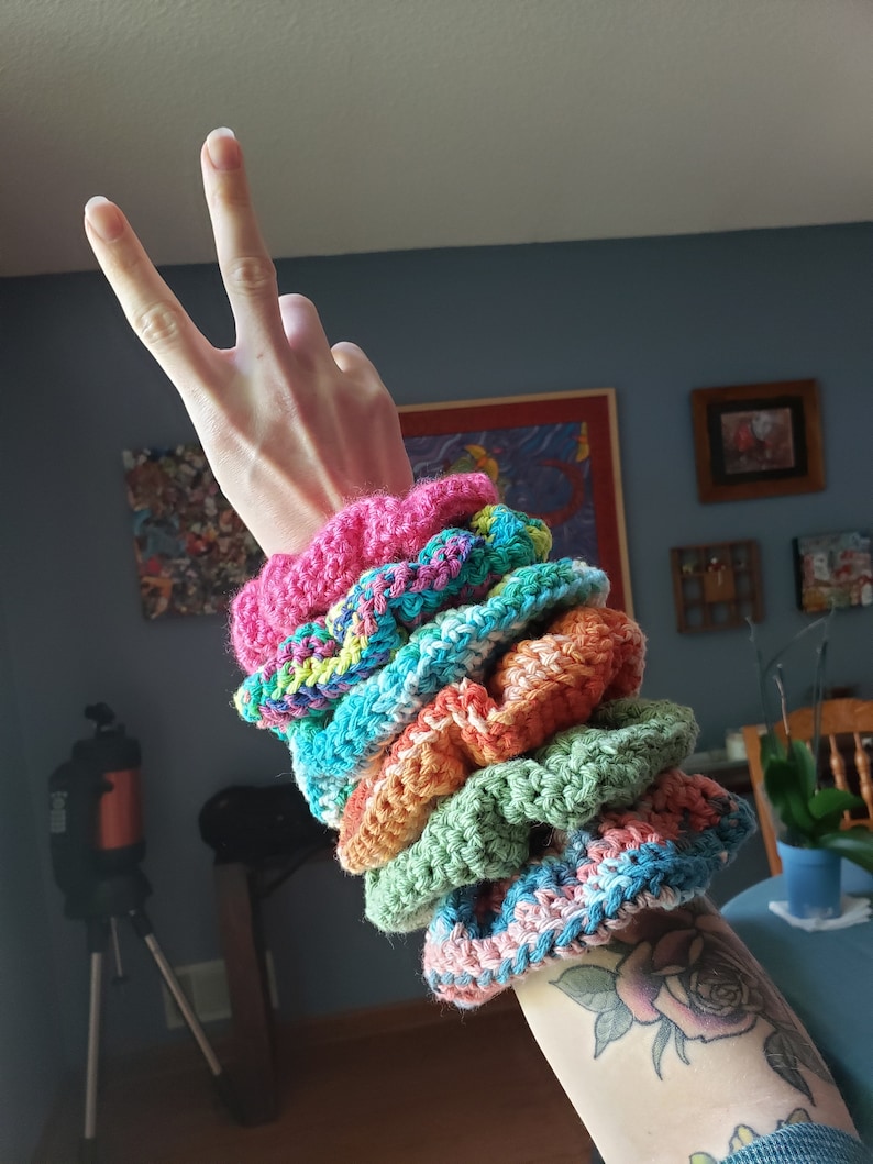 May include: A stack of six colorful crocheted hair scrunchies in pink, green, blue, orange, and teal.