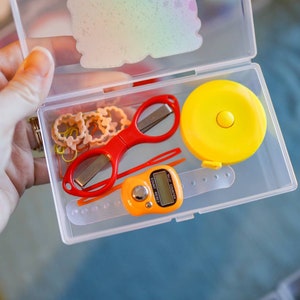 Pode incluir: Uma caixa de armazenamento de pl&aacute;stico transparente com uma variedade de pequenos itens, incluindo uma tesoura vermelha, uma fita m&eacute;trica amarela, um pequeno contador digital laranja e v&aacute;rios el&aacute;sticos de cabelo coloridos.