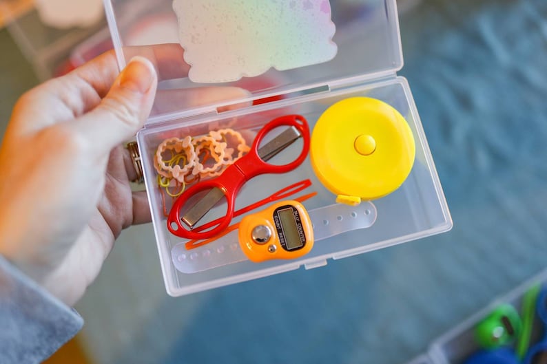 Pode incluir: Uma caixa de armazenamento de pl&aacute;stico transparente com uma variedade de pequenos itens, incluindo uma tesoura vermelha, uma fita m&eacute;trica amarela, um pequeno contador digital laranja e v&aacute;rios el&aacute;sticos de cabelo coloridos.