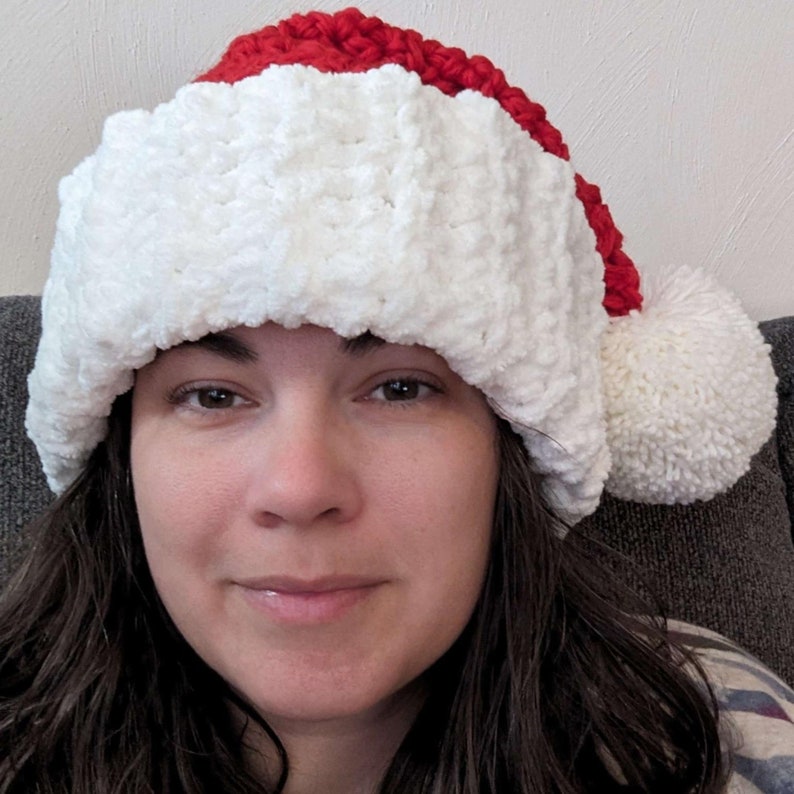 Op de afbeelding: Een witte en rode gehaakte kerstmanmuts met een witte pompon bovenop. De muts wordt gedragen door een persoon met lang bruin haar.