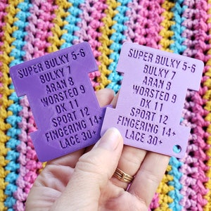 Op de afbeelding: Twee paarse garen gewichtmeters met tekst die de garen gewichten aangeeft, gehouden tegen een kleurrijke gehaakte achtergrond. De meters vermelden garen gewichten zoals Super Bulky, Bulky, Aran, Worsted, DK, Sport, Fingering en Lace.