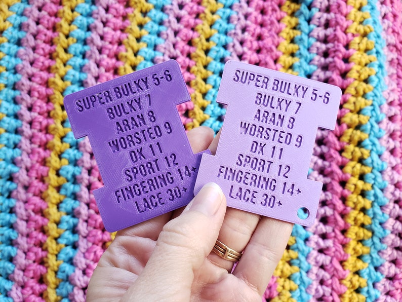 Op de afbeelding: Twee paarse garen gewichtmeters met tekst die de garen gewichten aangeeft, gehouden tegen een kleurrijke gehaakte achtergrond. De meters vermelden garen gewichten zoals Super Bulky, Bulky, Aran, Worsted, DK, Sport, Fingering en Lace.