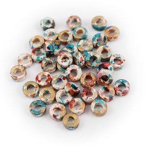 Puede incluir: Una colección de cuentas de cerámica de color donut con salpicaduras de pintura blanca, roja, dorada y azul. Las cuentas están dispuestas en una pila sobre un fondo blanco.