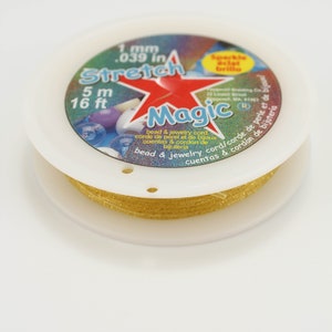 May include: A spool of gold-coloured Stretch Magic bead and jewellery cord. The label features a red star, the text 1 mm .039 in, and 5 m 16 ft. The cord is wound around a white plastic spool with a circular label on top with multi-coloured accents.