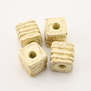 Op de afbeelding: Vier vierkante kralen in crème- en bruintinten. Elke kraal heeft een centraal gat en een getextureerd, gelaagd ontwerp. Deze kralen zijn waarschijnlijk bedoeld voor het maken van sieraden of knutselen. Ze meten ongeveer 1,5 cm vierkant.