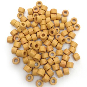 Könnte beinhalten: Ein Haufen kleiner, zylindrischer Perlen in einem warmen, hellbraunen Farbton. Jede Perle hat ein Loch in der Mitte, wodurch eine Donut-Form entsteht. Die Perlen sind auf einer weißen Oberfläche verstreut, ideal für Schmuckherstellung oder Bastelarbeiten.