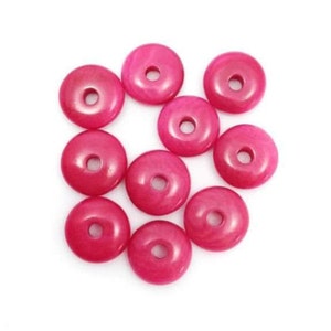 Könnte beinhalten: Neun fuchsiafarbene, donut-förmige Perlen mit glänzendem Finish sind auf weißem Hintergrund verstreut. Jede Perle hat ein zentrales Loch. Die Perlen sind gleichmäßig verteilt und zeigen ihre runde Form und lebendige Farbe.