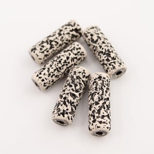 Può includere: Cinque perline cilindriche con un motivo maculato bianco e nero. Le perline sono disposte su una superficie bianca, mettendo in mostra il loro design strutturato. Ogni perlina ha un foro che attraversa il centro, suggerendo che sono per essere infilate.