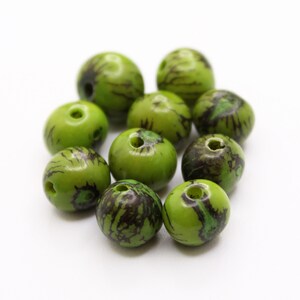 Pode incluir: Uma coleção de contas verdes com marmoreio castanho escuro e preto. Cada conta tem um buraco no centro e estão dispostas numa superfície branca. As contas têm um acabamento brilhante e uma forma redonda.