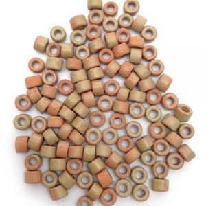 Pode incluir: Uma coleção de pequenas contas de cerâmica em tons de bege e laranja. As contas são cilíndricas e em forma de anel, com um furo no centro. Estão espalhadas numa superfície branca, adequadas para fazer joias ou artesanato.