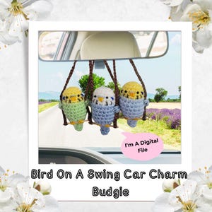 May include: Three crocheted budgie car charms hanging from a car's rearview mirror. The birds are in green, blue, and yellow, perched on a brown swing. The text "Bird On A Swing Car Charm Budgie" is at the bottom.