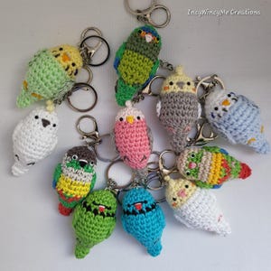 May include: A collection of 11 crocheted parakeet keyrings in various colours, including green, blue, pink, white, and grey. Each keyring features a small crocheted parakeet with a key ring attached.