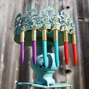 May include: Five pairs of bullet casing earrings, each painted a different color: purple, turquoise, red, and two shades of purple. The earrings are displayed on a turquoise stand.