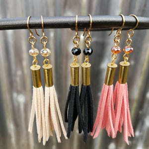 May include: Three pairs of dangle earrings with gold bullet casings, suede tassels, and crystal beads. The tassels are beige, black, and pink.