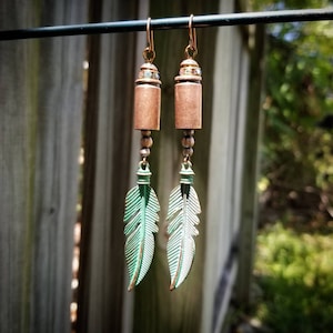 May include: A pair of dangle earrings featuring a copper bullet casing with rhinestones and a green feather charm.