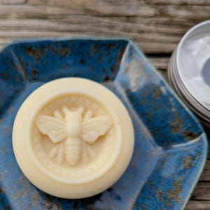 Op de afbeelding: Een ronde, witte, vaste zeep met een bijenontwerp op een blauw keramiek bord. De zeep zit in een container met een wit label waarop staat "Handmade wrapped in paper to preserve enjoy"