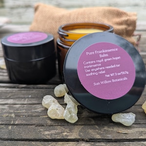 Puede incluir: Un frasco negro de bálsamo de incienso puro con una etiqueta morada, junto a un frasco con un bálsamo amarillo. La etiqueta dice "Contiene incienso hojan verde real. Usar donde sea necesario para aliviar. Peso neto 2 oz/56,7 g".