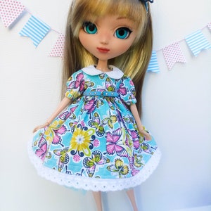 May include: A doll with long blonde hair wearing a blue dress with a white collar and a butterfly print. The dress has a white lace trim and a blue belt with silver studs. The doll is wearing a blue headband with a butterfly on it.