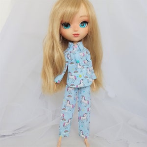 Könnte beinhalten: Eine Puppe trägt einen blauen Schlafanzug mit einem Einhorn-Druck. Der Schlafanzug hat lange Ärmel und lange Hosen. Die Puppe hat blonde Haare und blaue Augen.