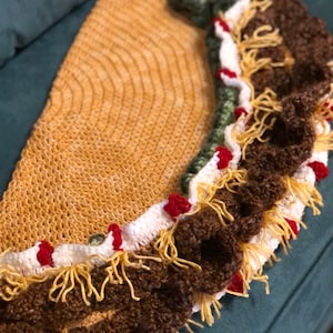 Puede incluir: Una manta de ganchillo con forma de taco, de hilo marrón, amarillo, blanco, rojo y verde. La manta tiene un borde rizado y flecos.