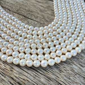May include: Close-up of multiple strands of lustrous, creamy white pearls. The pearls are round and evenly sized, creating a smooth, elegant texture. The background is a weathered wooden surface, providing a rustic contrast to the pearls.