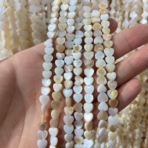 Peut inclure: Gros plan sur des rangées de perles en forme de cœur, blanches et crème. Les perles sont enfilées, créant une esthétique délicate. Les perles sont faites d'un matériau nacré, reflétant la lumière. Idéal pour la création de bijoux.