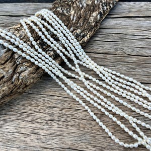 May include: Several strands of white, oval-shaped pearls cascade over a piece of weathered wood. The pearls are strung together, creating a delicate and elegant appearance. The background is a rustic wooden surface.